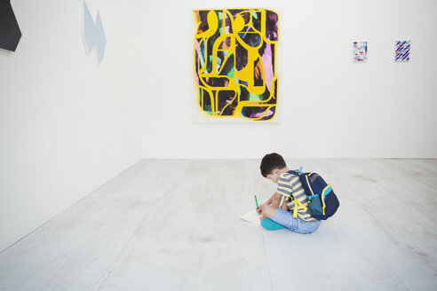 Junge mit kurzen schwarzen Haaren, der einen Rucksack trägt, sitzt in einer Kunstgalerie mit Stift und Papier auf dem Boden und betrachtet ein modernes Gemälde. - MINF06850