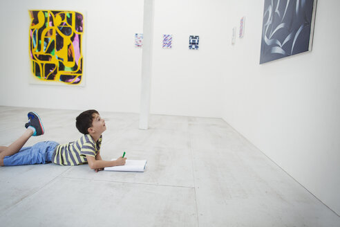 Junge mit kurzen schwarzen Haaren liegt in einer Kunstgalerie mit Stift und Papier auf dem Boden und betrachtet ein modernes Gemälde. - MINF06851