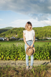 A young woman holding a sun hat, standing on a road by open fields of rice paddies. - MINF06877