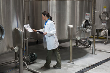 Caucasian female technician checking large stainless steel processing tanks in a bottling plant. that makes flavoured bottled water. - MINF06890