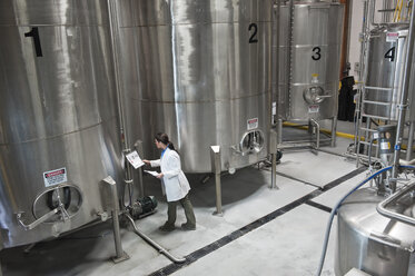 Caucasian female technician checking large stainless steel processing tanks in a bottling plant. that makes flavoured bottled water. - MINF06893