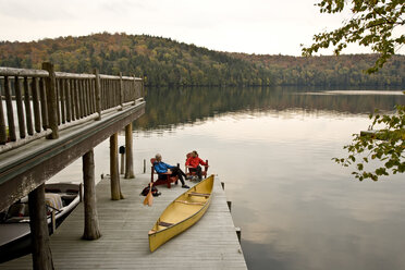 Young couple sitting on dock with canoe in early morning, Lake Placid, New York, USA. - AURF00044