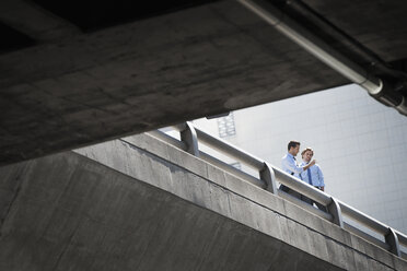 Two businessmen standing leaning on a railing on a city walkway, view from below. - MINF06977