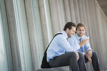 Zwei Geschäftsleute in Hemd und Krawatte sitzen vor einem Bürogebäude und überprüfen ihre Smartphones. - MINF06982
