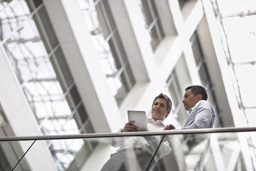 Two men, business colleagues, standing by a railing in an atrium or courtyard, looking at a digital tablet. - MINF07019