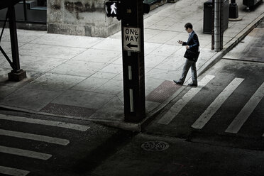 Ein Mann geht über eine Straßenkreuzung und schaut auf ein Telefon in seiner Hand. - MINF07031