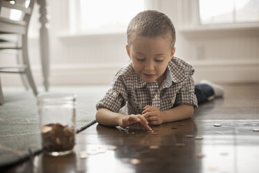 A child lying on his stomach on the floor playing with coins and putting them in a glass jar. - MINF07033