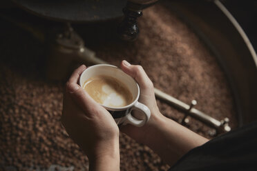 Eine Person hält eine frisch gebrühte Tasse über einer Metalltrommel mit gerösteten Kaffeebohnen. - MINF07098