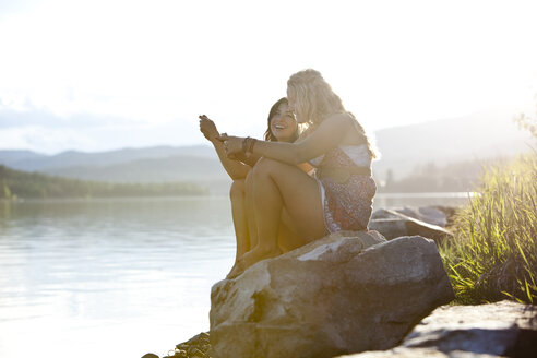 Zwei junge Frauen lachen, während sie bei Sonnenuntergang am See eine SMS schreiben. - AURF00118