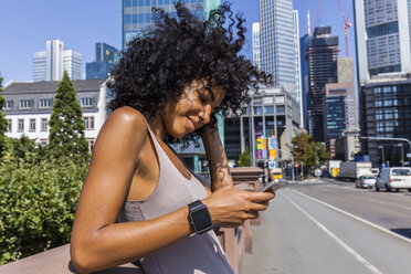 Germany, Frankfurt, portrait of smiling young woman with curly hair using cell phone in the city - TCF05578