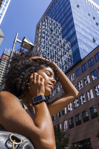 Deutschland, Frankfurt, Porträt einer jungen Frau mit Smartwatch am Telefon vor einem Wolkenkratzer, lizenzfreies Stockfoto