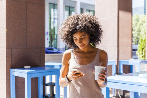 Lächelnde junge Frau mit Kaffee zum Mitnehmen schaut auf ihr Handy, lizenzfreies Stockfoto