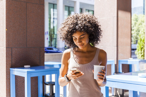 Lächelnde junge Frau mit Kaffee zum Mitnehmen schaut auf ihr Handy - TCF05593