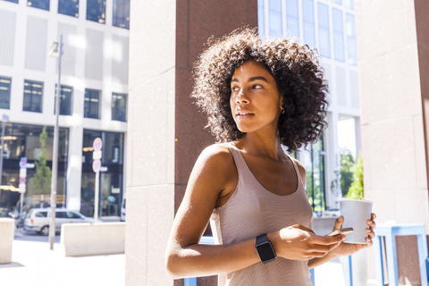 Porträt einer jungen Frau mit Kaffee zum Mitnehmen und Handy, die etwas beobachtet, lizenzfreies Stockfoto