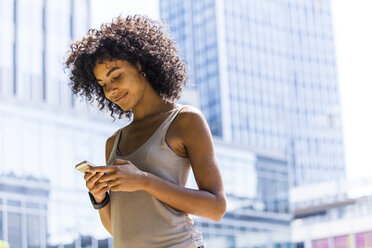 Germany, Frankfurt, smiling young woman with curly hair looking at cell phone in front of skyscrapers - TCF05614