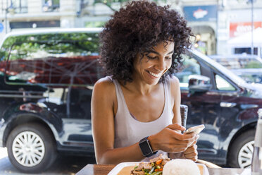 Porträt einer lächelnden jungen Frau, die auf dem Bürgersteig sitzt und auf ihr Mobiltelefon schaut - TCF05615