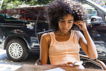 Porträt einer jungen Frau mit lockigem Haar, die in einem Straßencafé sitzt und auf ihr Mobiltelefon schaut - TCF05617
