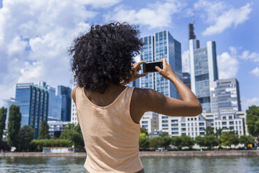 Germany, Frankfurt, back view of young woman with curly hair taking photo with smartphone - TCF05623