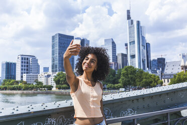 Germany, Frankfurt, portrait of content young woman with curly hair taking selfie in front of skyline - TCF05624