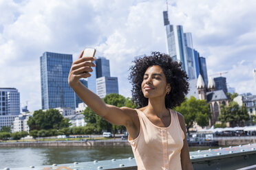 Germany, Frankfurt, portrait of content young woman with curly hair taking selfie in front of skyline - TCF05625