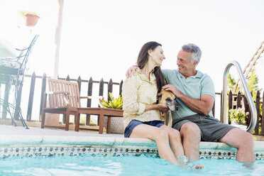 A couple sitting on the edge of a swimming pool, with their dog between them. - MINF07160