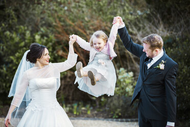 Braut und Bräutigam, ein Paar an seinem Hochzeitstag, das ein kleines Mädchen zwischen sich in der Luft schwingt. - MINF07199