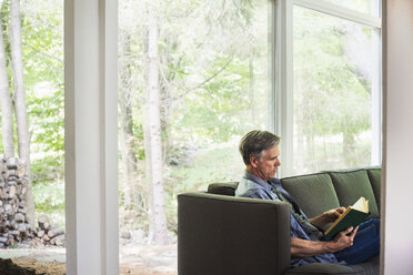 Ein Mann sitzt am Fenster auf einem Sofa und liest ein Buch. - MINF07227