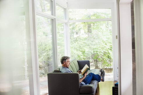 Ein Mann sitzt am Fenster auf einem Sofa und liest ein Buch. - MINF07228