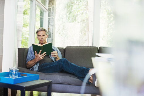 Ein Mann sitzt am Fenster auf einem Sofa und liest ein Buch. - MINF07229