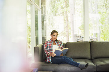 A woman sitting on a sofa by a window, reading a book. - MINF07230