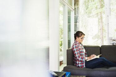 A woman sitting on a sofa by a window, reading a book. - MINF07231