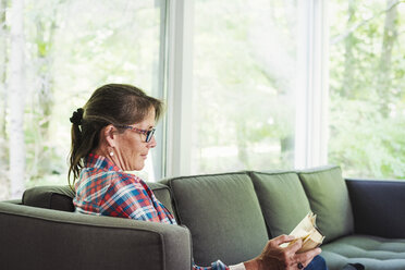 Eine Frau in einem karierten Hemd sitzt auf einem Sofa und liest ein Buch. - MINF07232