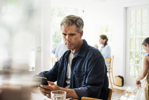 Mann sitzt in einem Café und schaut auf sein Smartphone. Unscharfer Vordergrund. - MINF07257