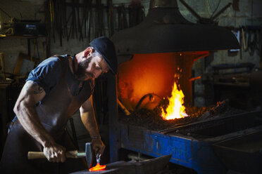 A blacksmith strikes a pieces of red hot metal on an anvil with a hammer in a workshop. - MINF07278