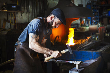 A blacksmith uses complex tools to hammer a cone of red hot metal on an anvil in a workshop. - MINF07279