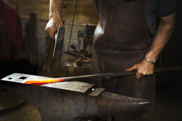 Ein Schmied schlägt in einer Werkstatt mit einem Hammer ein Stück glühendes Metall auf einen Amboss. - MINF07282