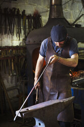 A blacksmith in a leather apron holds a thin length of metal against an anvil in a workshop. - MINF07285