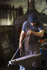 Ein Schmied in einer Lederschürze hält ein dünnes Metallstück gegen einen Amboss in einer Werkstatt. - MINF07285