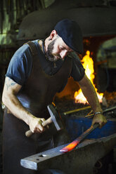 A blacksmith strikes a length of red hot metal on an anvil with a hammer in a workshop. - MINF07289