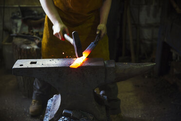 A blacksmith strikes a length of red hot metal on an anvil with a hammer in a workshop. - MINF07293