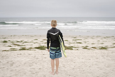 Blond boy standing on a sandy beach, holding a bodyboard. - MINF07326