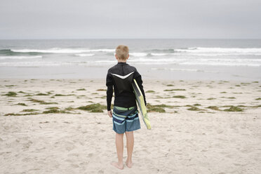 Ein blonder Junge steht am Sandstrand und hält ein Bodyboard. - MINF07326