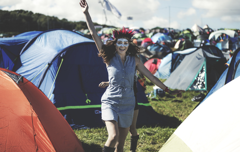 Junge Frau auf einem Sommermusikfestival, Gesicht bemalt, mit Federschmuck, steht in der Nähe des von Zelten umgebenen Campingplatzes, den Arm erhoben, lächelnd., lizenzfreies Stockfoto