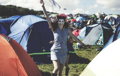 Junge Frau auf einem Sommermusikfestival, Gesicht bemalt, mit Federschmuck, steht in der Nähe des von Zelten umgebenen Campingplatzes, den Arm erhoben, lächelnd. - MINF07625