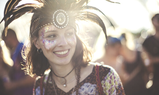 Junge Frau auf einem Sommermusikfestival mit Federkopfschmuck und geschminktem Gesicht, lächelnd. - MINF07629