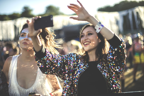 Junge lächelnde Frau auf einem Sommermusikfestival, die eine bunte Paillettenjacke trägt und ein Foto mit ihrem Smartphone macht., lizenzfreies Stockfoto