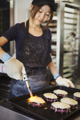 Frau arbeitet in einer Bäckerei, trägt Ofenhandschuhe, benutzt eine Lötlampe und schmilzt Käse auf Sandwiches. - MINF07692