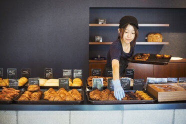 Eine Frau arbeitet in einer Bäckerei und legt frisch gebackene Croissants und Kuchen auf großen Tabletts auf einen Tresen. - MINF07693