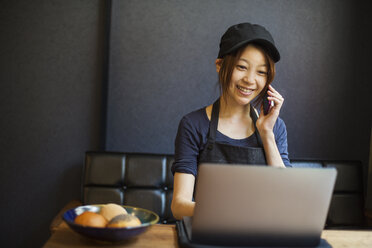 Frau arbeitet in einer Bäckerei, trägt eine Baseballmütze, sitzt am Tisch vor einem Laptop und benutzt ein Mobiltelefon. - MINF07694