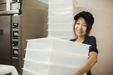 Woman working in a bakery, wearing baseball cap, carrying stack of white plastic crates. - MINF07695